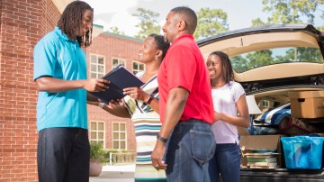 African descent teenage boy moves into the college dorm after high school. He is excited about his next new adventure. His family says goodbye outside the campus dorm. Vehicle filled with his belongings is in background.