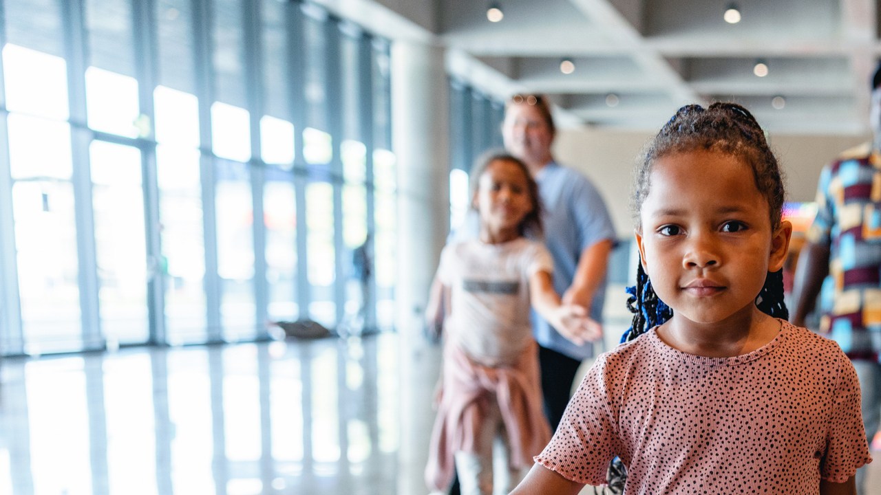 Three-quarter front shot with blurred background of a mixed-race little kid taking the lead and walking in front of their multicultural family to be the first one who finds the exit of the airport.