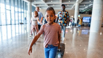 Three-quarter front shot with blurred background of a mixed-race little kid taking the lead and walking in front of their multicultural family to be the first one who finds the exit of the airport.
