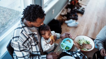 High angle view looking down at a cute multiracial toddler girl sitting in her father's lap and feeding herself while having dinner with her multi-generation family during a weekend gathering.