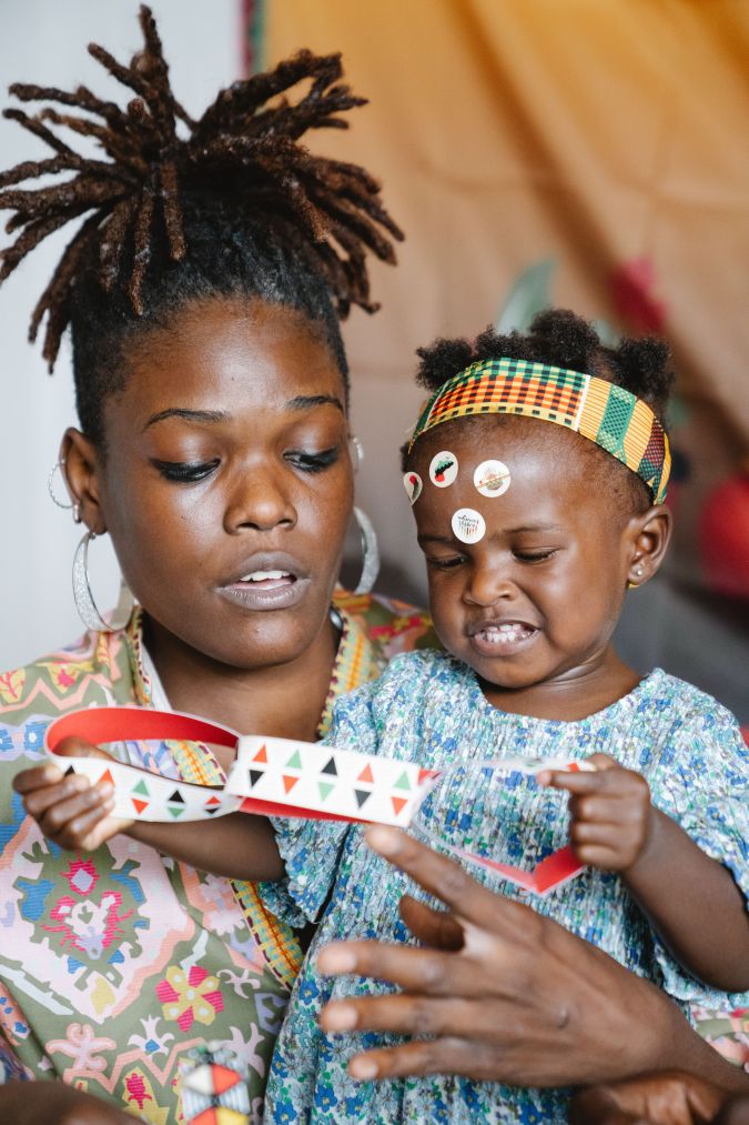 A woman and a young girl are engaged in a creative activity, with the woman showing a colorful paper strip to the girl, who has stickers on her forehead and looks focused.