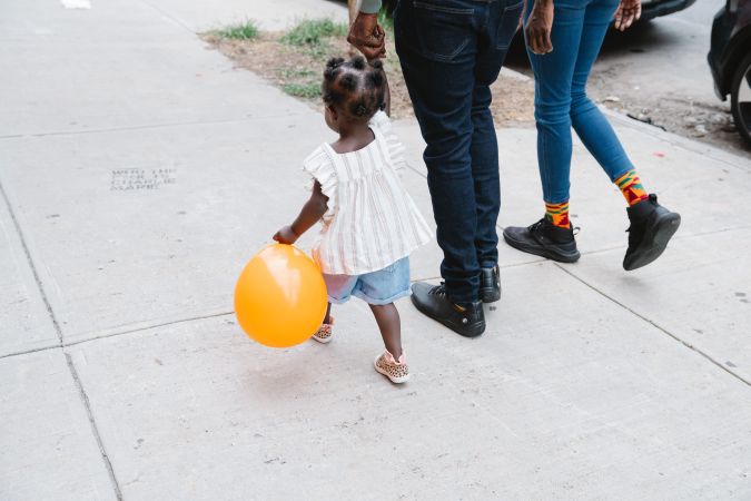 A young child holding an orange balloon walks alongside two adults on a sidewalk.