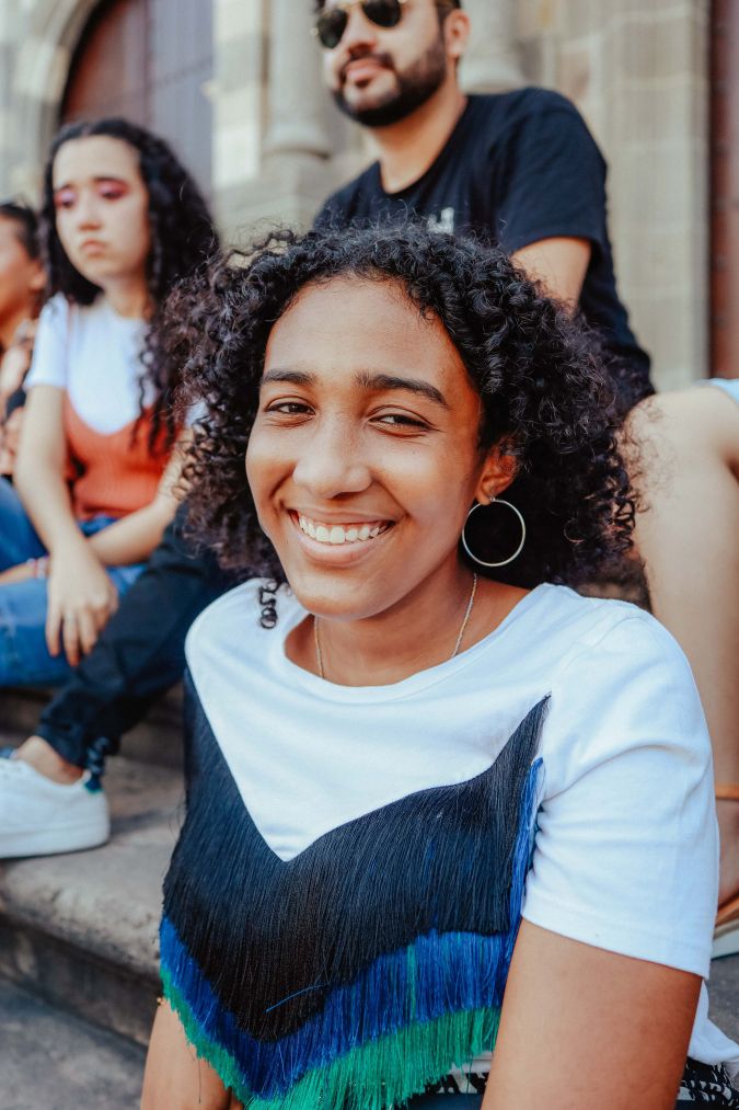 A smiling woman with curly hair wearing a white shirt with colorful fringe sits on steps, with friends in the background.