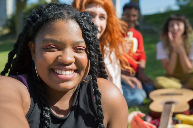A group of friends enjoying a sunny day outdoors, with one person smiling in the foreground while holding a camera. The background shows several others laughing and sitting on the grass, surrounded by snacks and a guitar.
