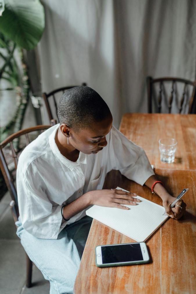 Person sitting at a wooden table, writing in a notebook with a pen, while a smartphone lies nearby.