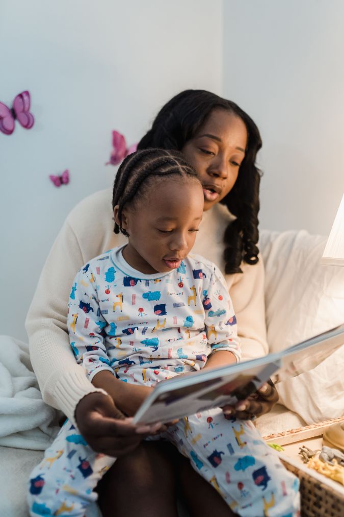 A mother reading a book to her child, who is sitting on her lap, both are cozy in a warmly lit room decorated with butterflies.