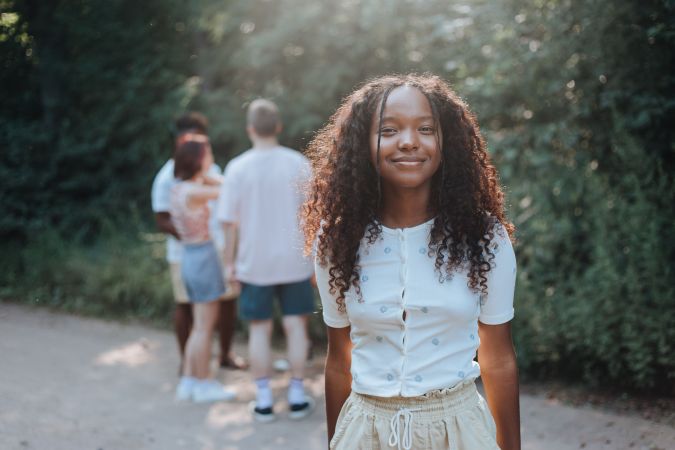 A young girl with curly hair smiles at the camera in a sunlit outdoor setting, while a small group of people stands in the background.