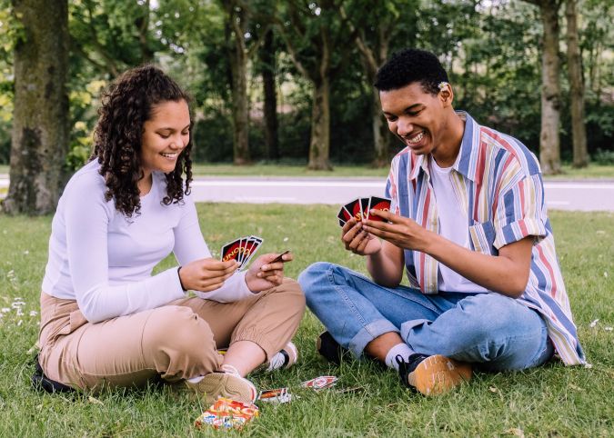 Two young people sitting on the grass, playing a card game and smiling at each other. One is holding cards while the other looks at their hand. The scene is set in a park with trees in the background.