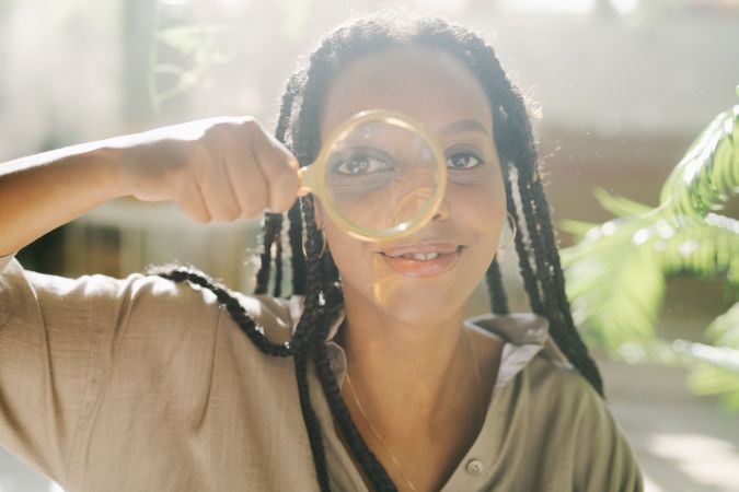 A woman with long braided hair holds a magnifying glass up to her eye, smiling playfully. Natural light fills the background, and there are green plants nearby.