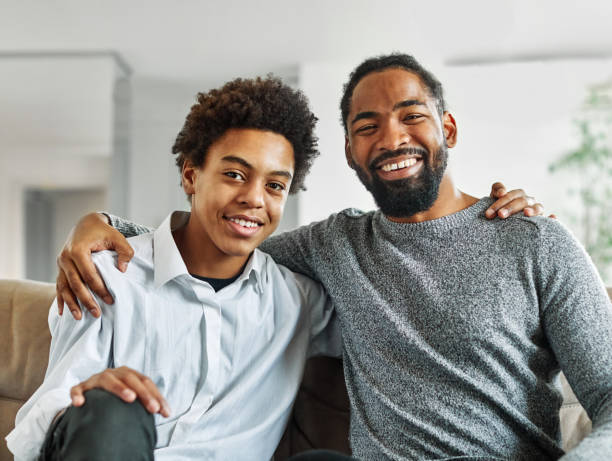 A teenage boy and an adult man smiling together while seated on a couch in a cozy living room.