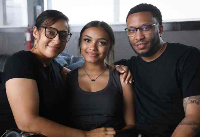 A young woman sits between two smiling adults, all wearing black shirts. They are posing together with friendly expressions in a casual indoor setting.