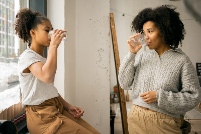 A young girl and a woman enjoy drinking water from glass cups while sitting indoors near a window.