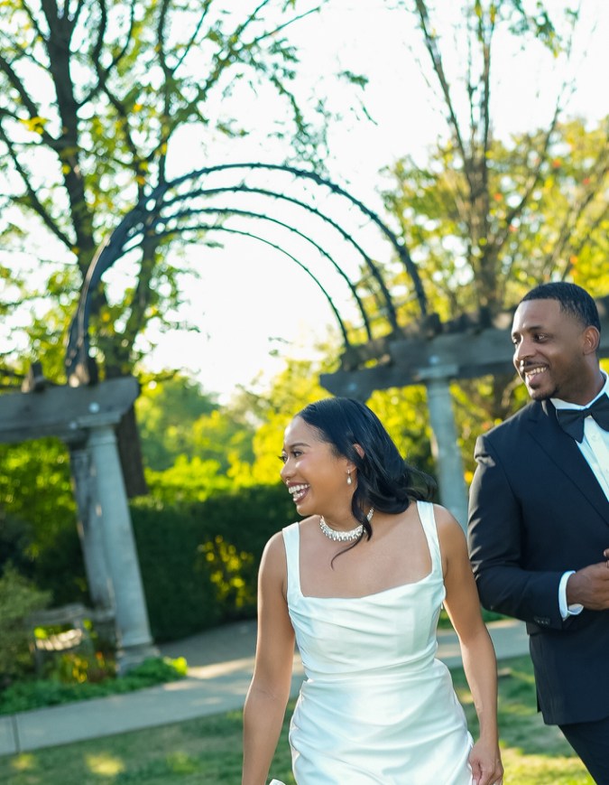A joyful couple walking hand in hand in a garden setting, with the woman wearing a sleek white dress and holding a bouquet of flowers, while the man is dressed in a black tuxedo.