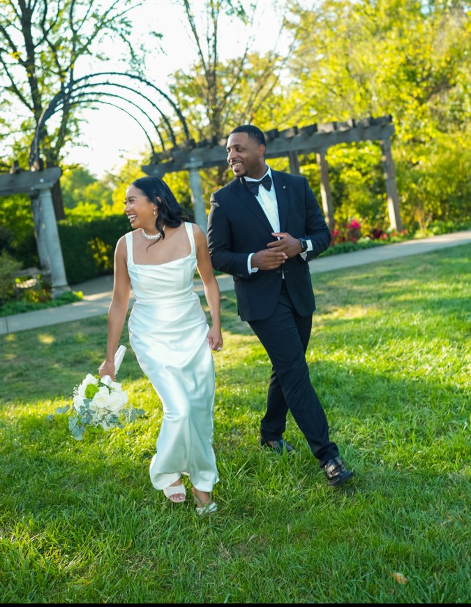 A joyful couple walking hand in hand in a garden setting, with the woman wearing a sleek white dress and holding a bouquet of flowers, while the man is dressed in a black tuxedo.
