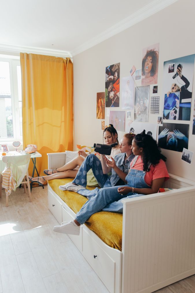 Three young women sitting on a white daybed in a brightly lit room, watching a video on a portable screen. The walls are decorated with various photos and posters, and a yellow curtain is partially drawn over the window.