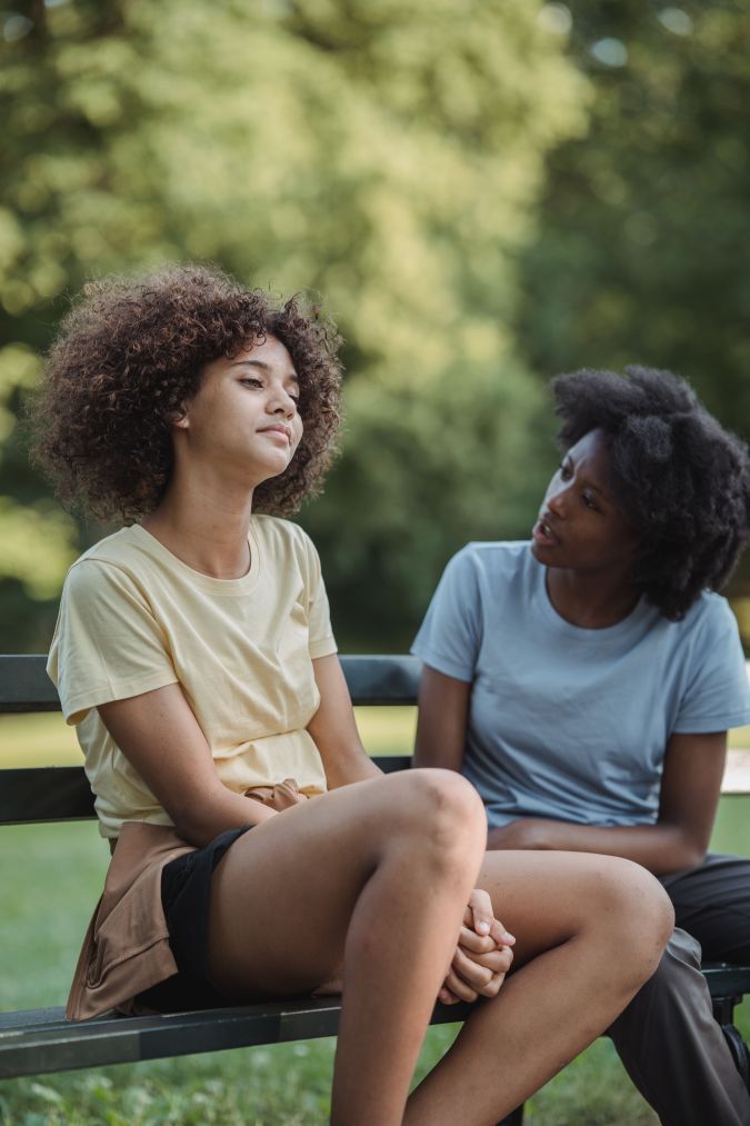 Two young women sit on a park bench, engaged in a serious conversation. One woman, wearing a yellow shirt, appears contemplative, while the other, in a blue shirt, looks attentively at her.