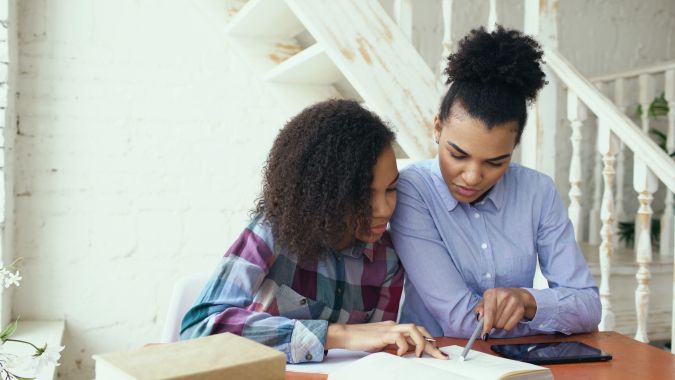 Two women studying together at a table with a notebook and a tablet, focusing on their work in a bright and cozy environment.