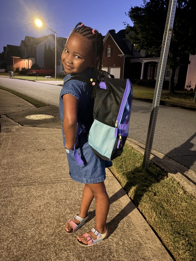 A young girl with braided hair is standing on a sidewalk at dusk, smiling while wearing a denim dress and a colorful backpack.