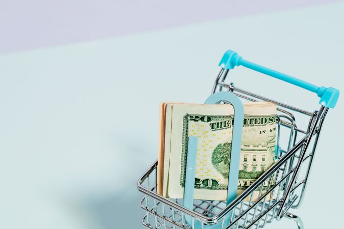 A miniature shopping cart filled with folded dollar bills, resting on a light blue background.