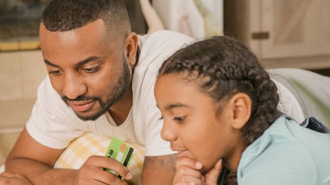 A man and a young girl lying on a bed, engaging with a credit card and looking at a screen together.