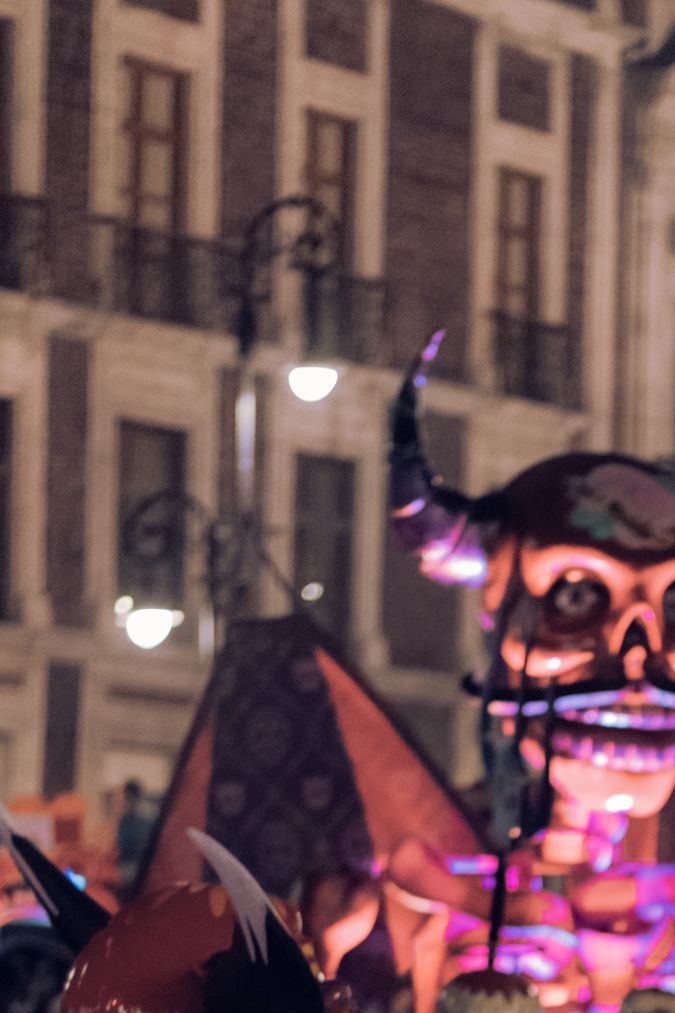 A festive nighttime parade scene featuring participants in elaborate costumes, including a figure in a shiny silver cape and helmet, alongside colorful decorations and large vibrant sculptures resembling skulls and mythical creatures.