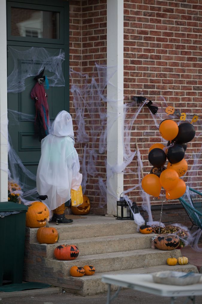 A child dressed in a ghost costume stands at a decorated front door with spider webs, a witch decoration, and pumpkins, while holding a candy bucket.