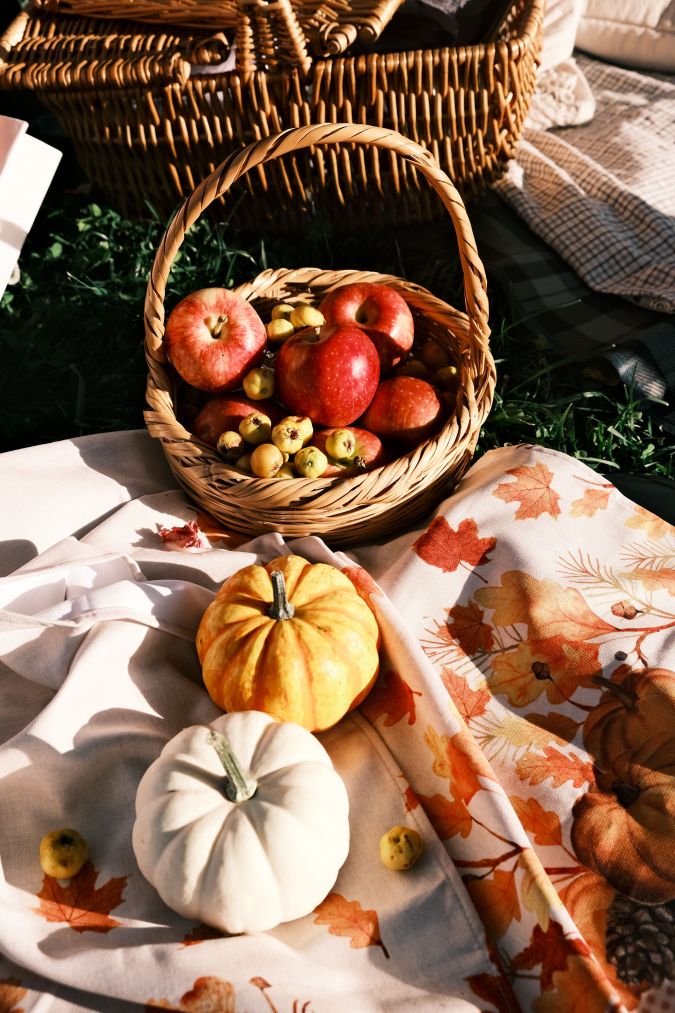 A basket filled with red apples and small fruits sits on a grass surface, accompanied by a white and an orange pumpkin on a patterned cloth with autumn leaves.
